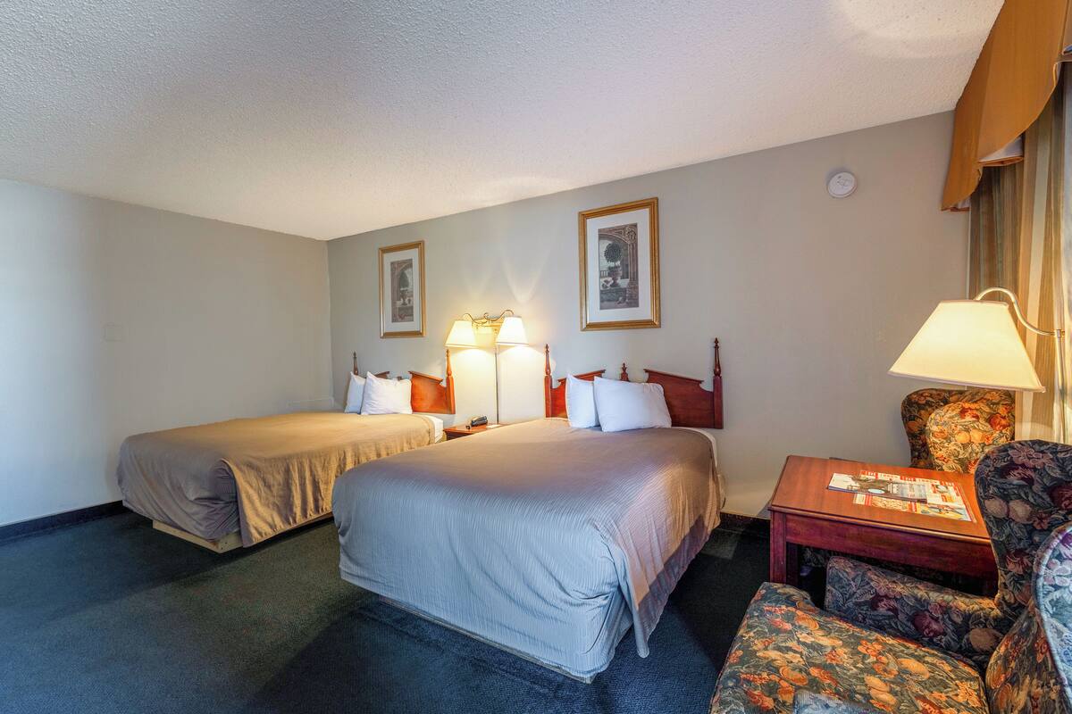 A hotel room with two beds, a desk, a chair, and a lamp.
