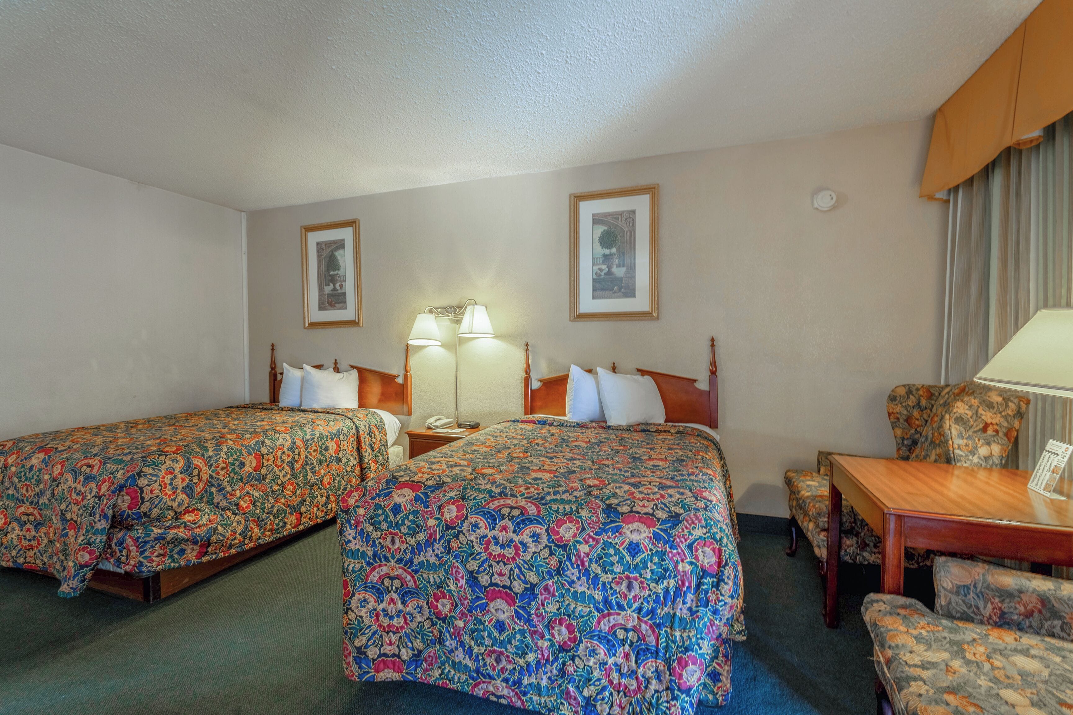 A hotel room with two beds, a desk, and a chair. There are two lamps on the desk and a telephone on the nightstand.