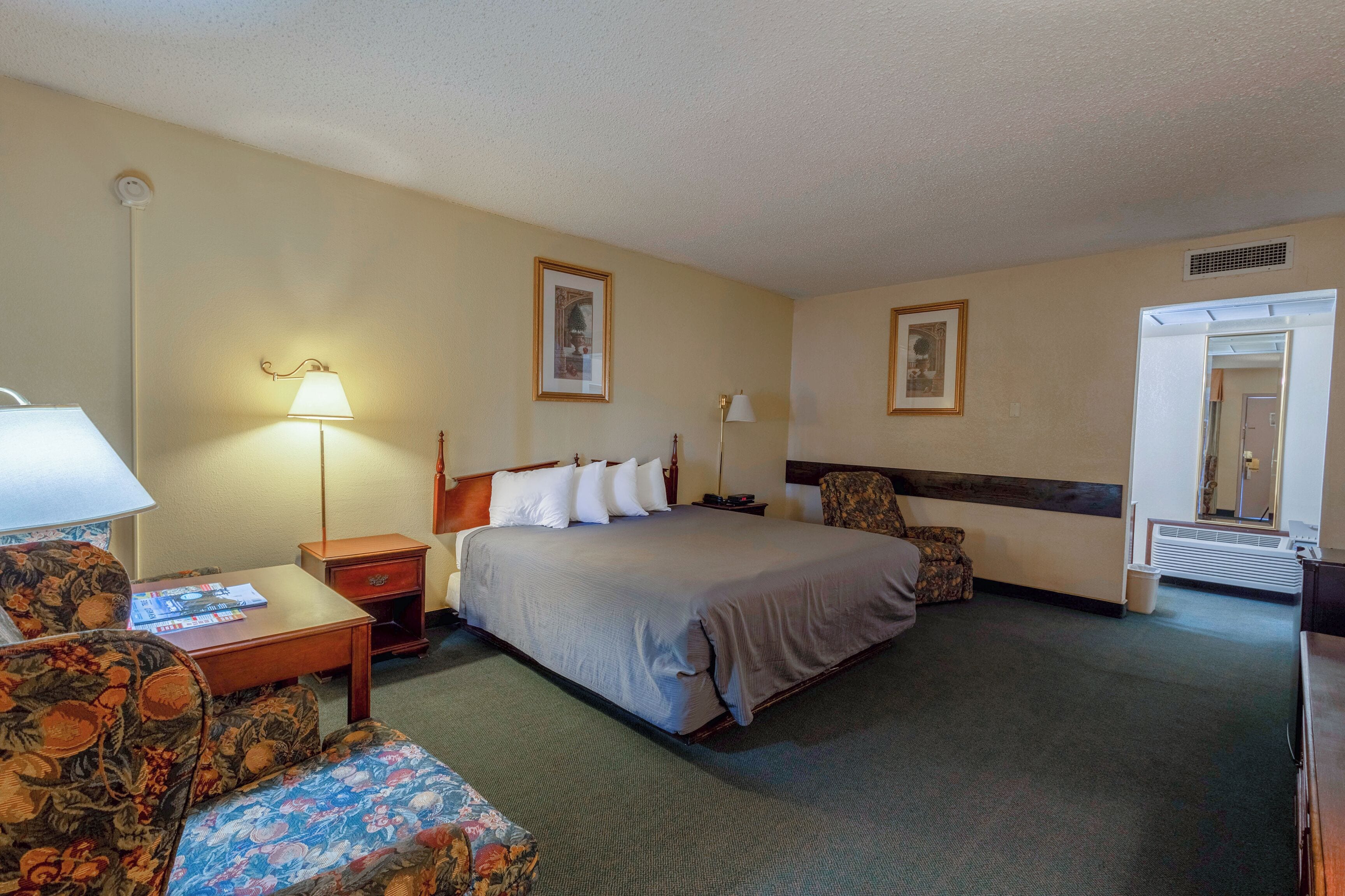 A hotel room with a bed, two chairs, a nightstand, a lamp, and a framed picture on the wall.