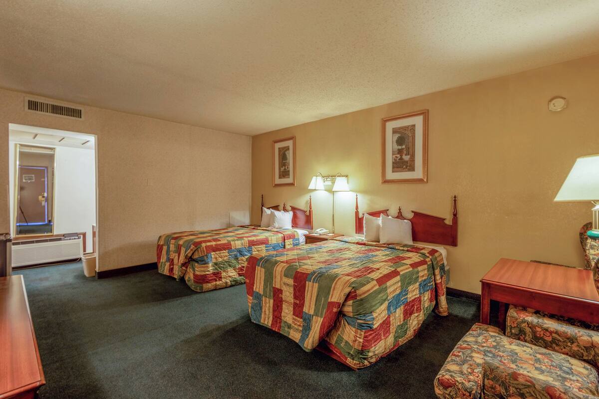 A hotel room with two beds, a desk, a chair, and a lamp.