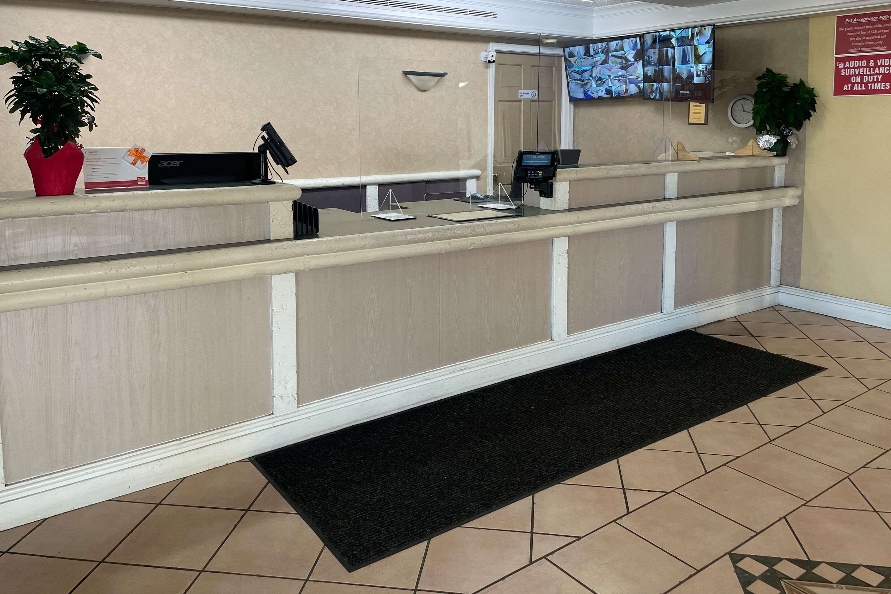 Een hotel receptie met een receptiebalie, een plant, een monitor die beveilingsbeelden toont en een zwarte mat op de vloer.