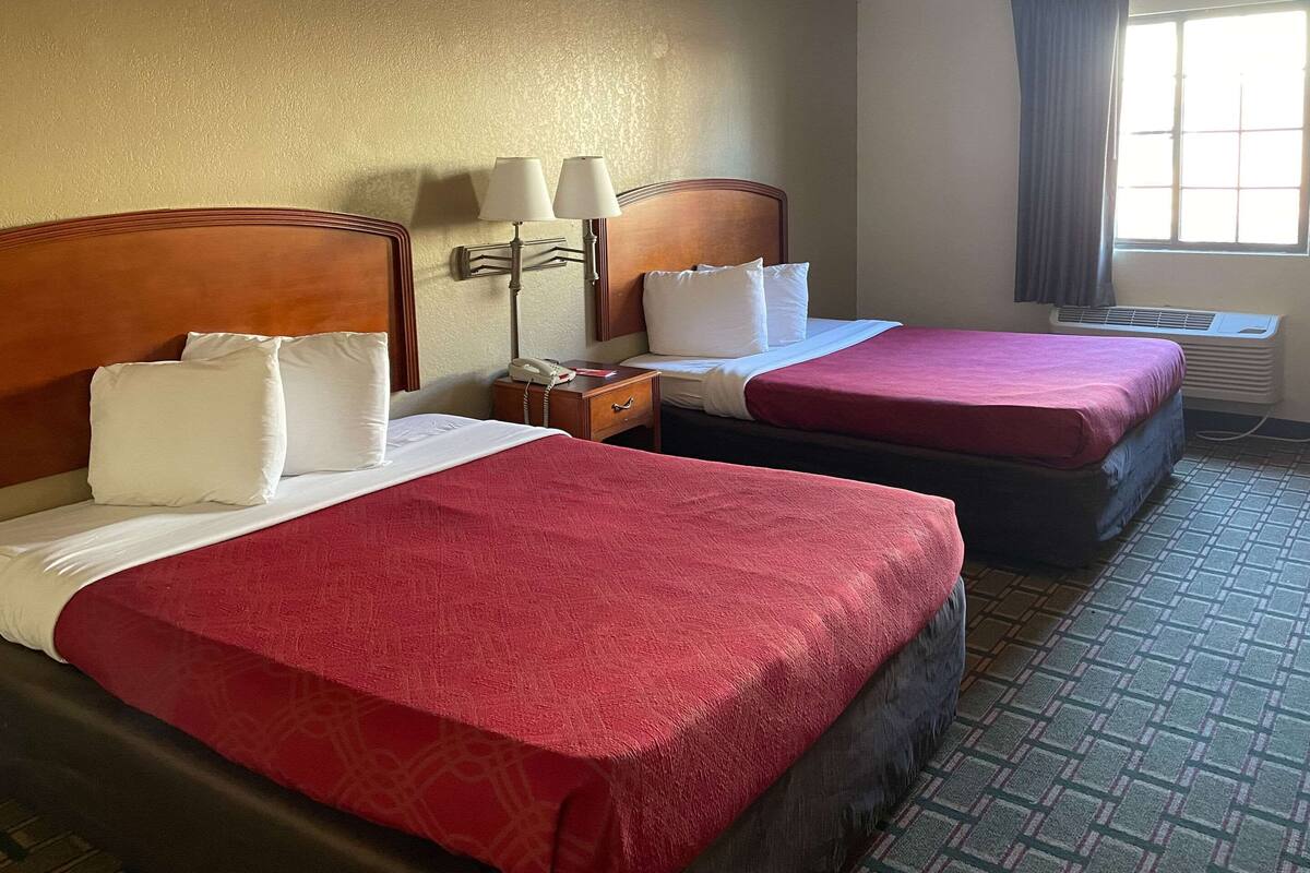 A hotel room with two beds, each with a red bedspread and white pillows. There is a nightstand with a lamp and a phone, and an air conditioning unit near the window.