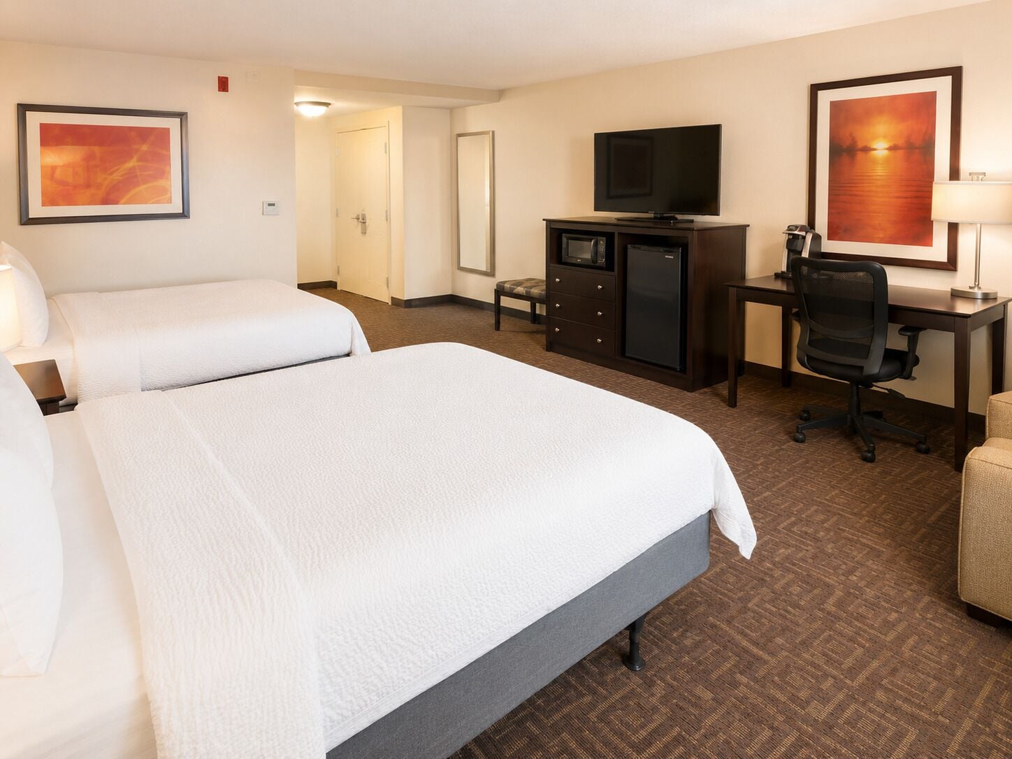 A hotel room with two beds, a desk with a computer, a TV, and artwork on the walls.