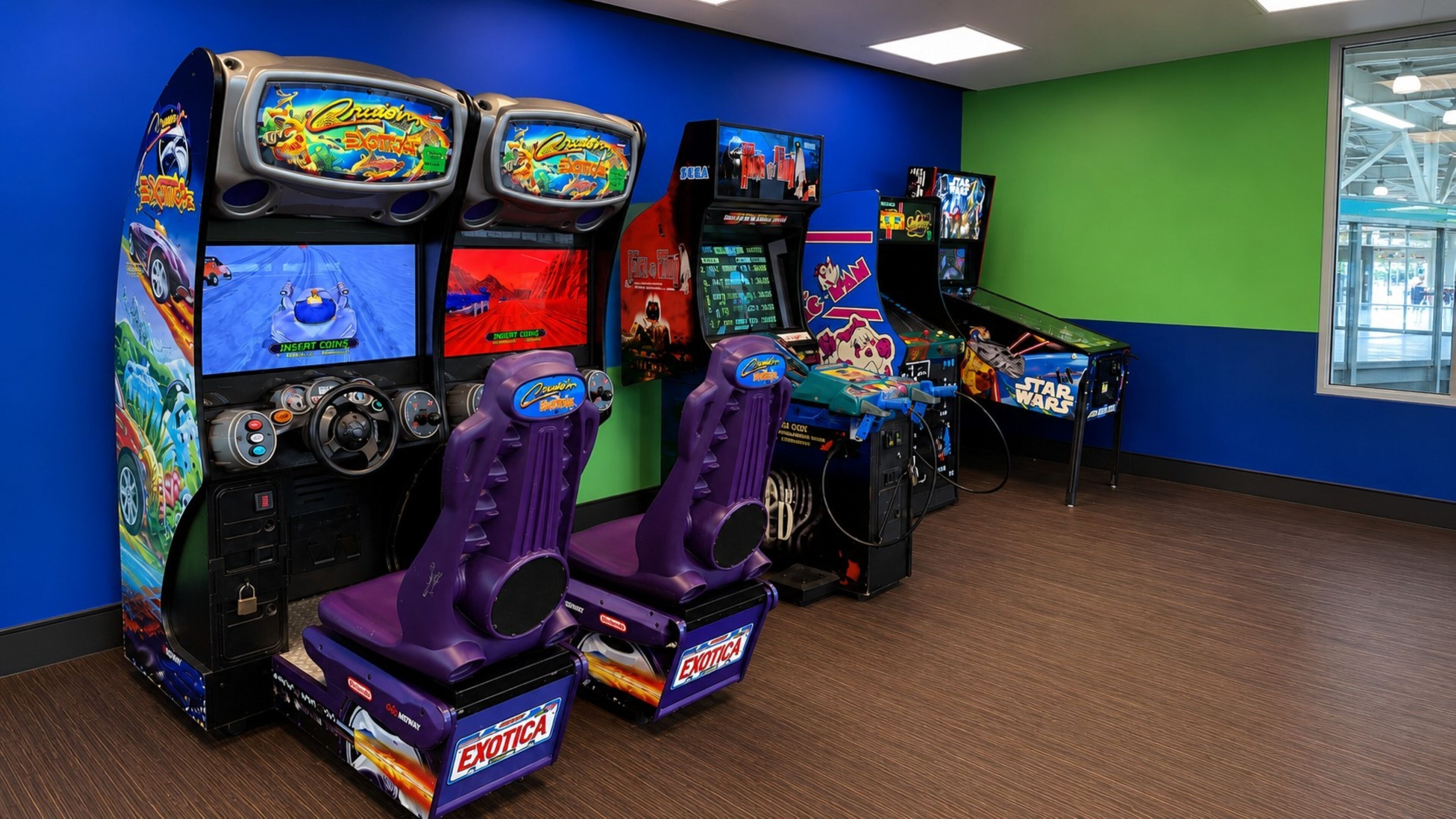 A row of arcade gaming machines with colorful graphics and multiple screens, set against a blue and green wall in a room with wooden flooring.