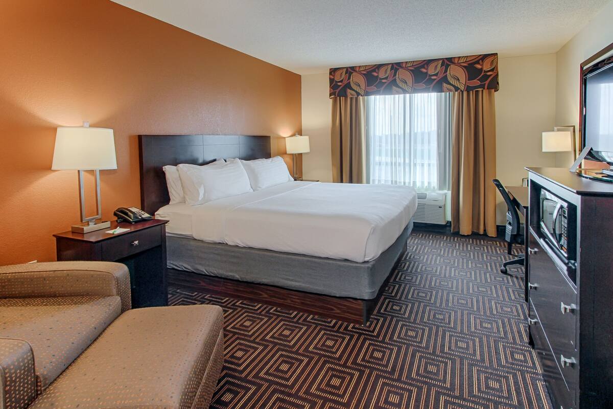 A hotel room with a large bed, two bedside tables with lamps, a desk with a computer, a chair, and a patterned carpet.