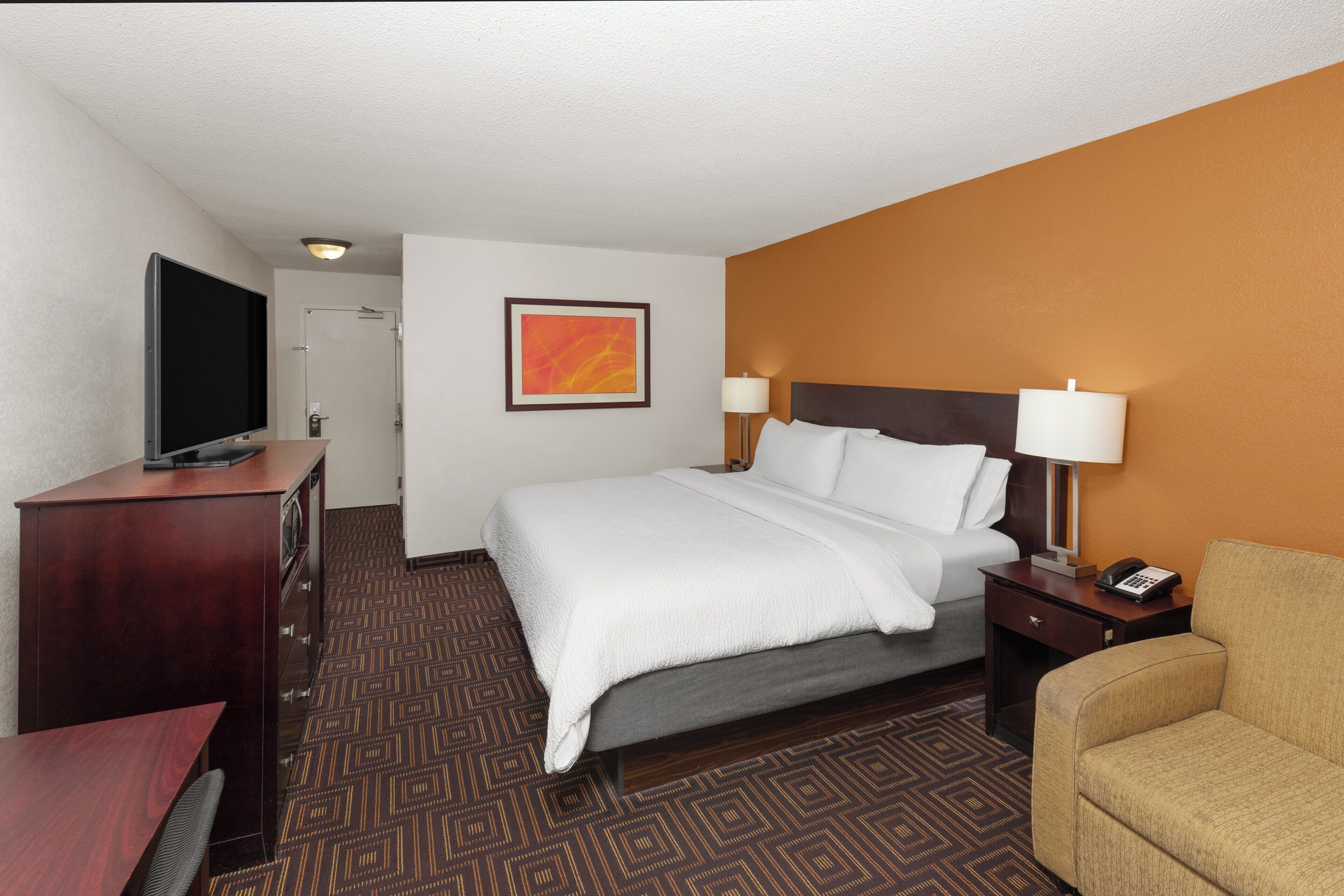 A hotel room with a bed, a desk with a television, a chair, a lamp, and a painting on the wall.