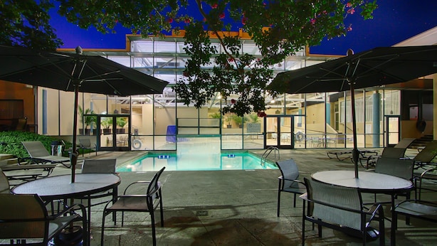Zona de piscina al aire libre con sillas de descanso y sombrillas, rodeada por un edificio con grandes ventanales.