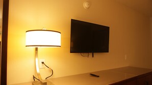 A hotel room with a flat-screen TV mounted on the wall, a bedside table with a lamp, and a remote control on the table. - All Seasons Inn & Suites (Memphis)