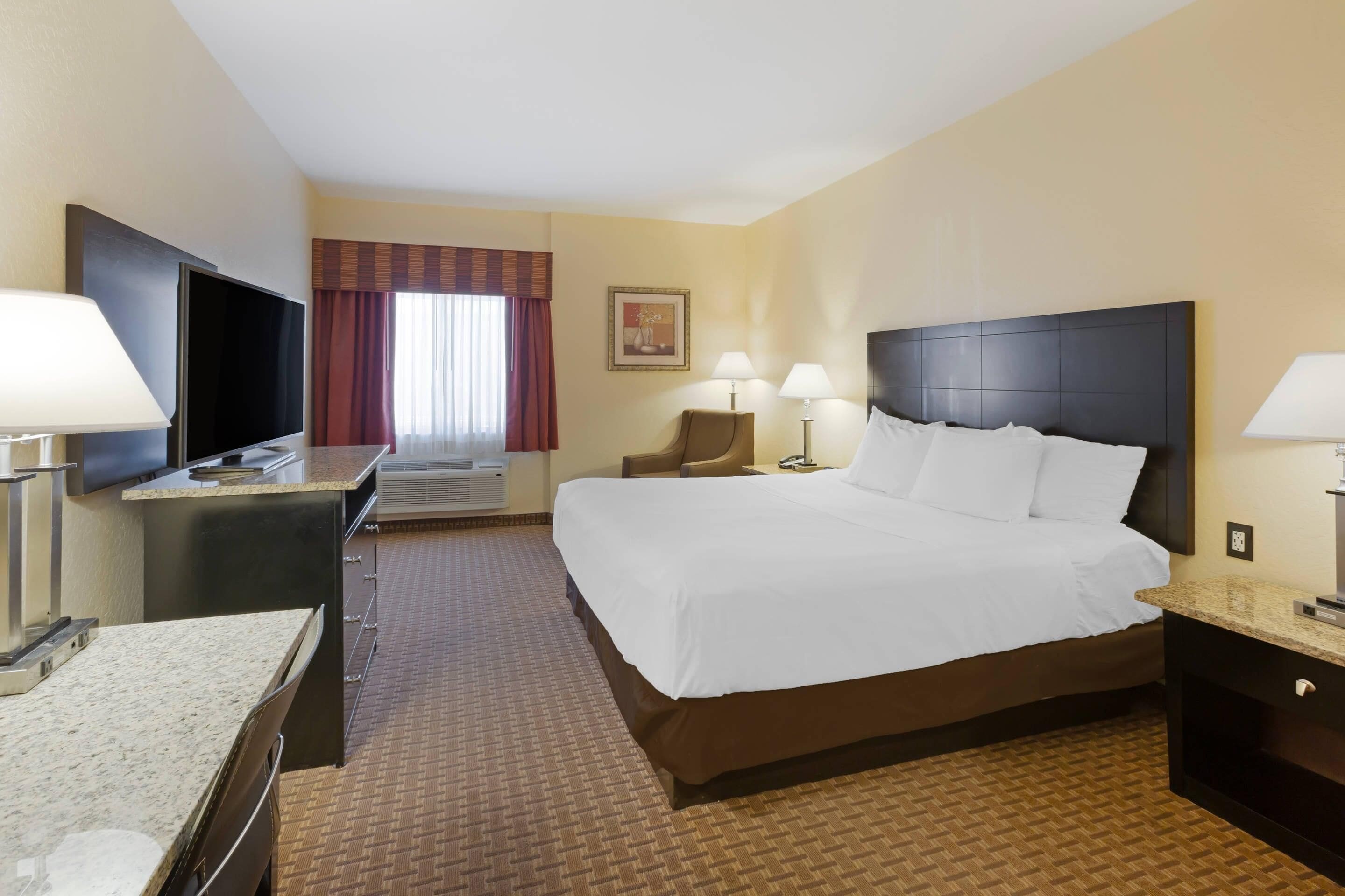 Una habitación de hotel con cama, mesitas de noche, un escritorio con televisor de pantalla plana, una silla, una ventana con cortinas y un cuadro en la pared.