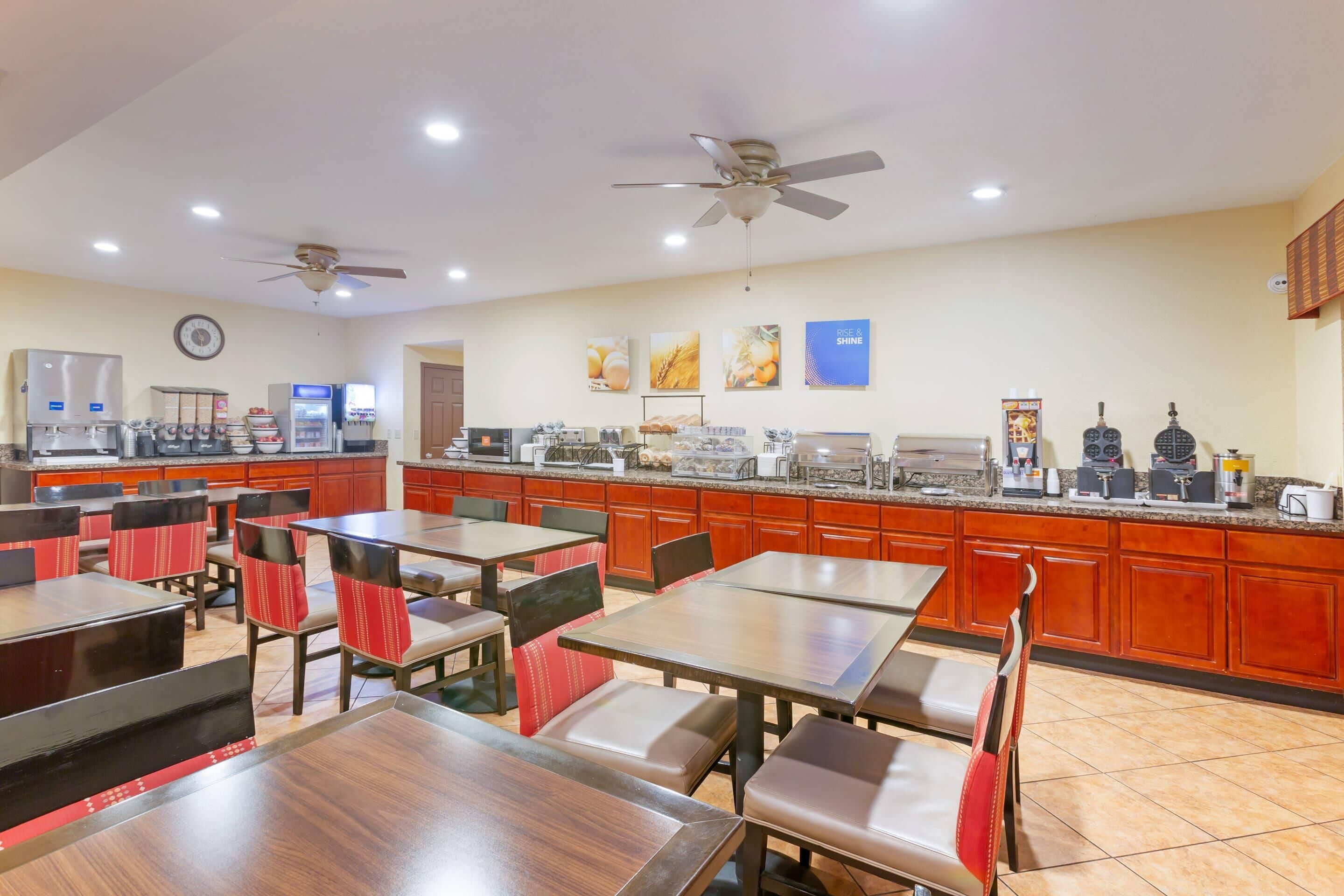 A hotel dining area with tables and chairs, a kitchen in the background, and ceiling fans.