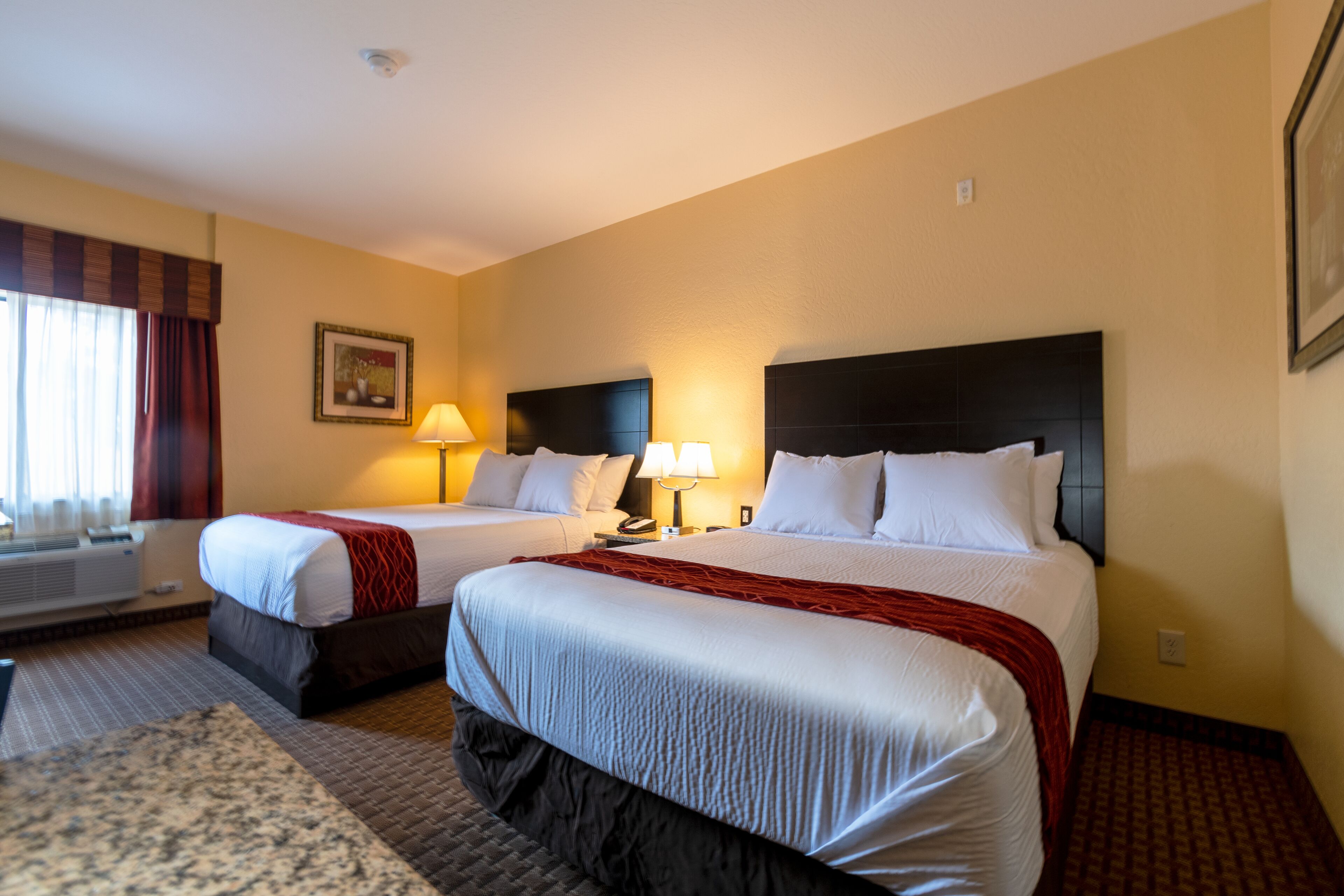A hotel room with two beds, a large window with curtains, a framed picture on the wall, and a carpeted floor.