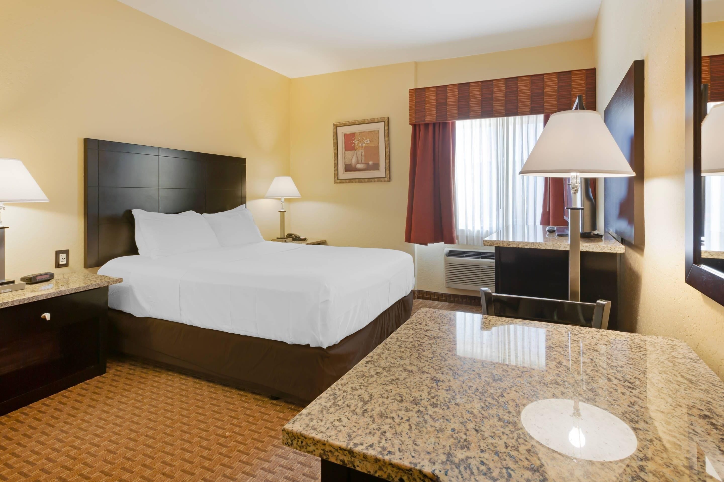 A hotel room with a bed, bedside lamps, a desk, a chair, a large mirror, and a window with curtains.