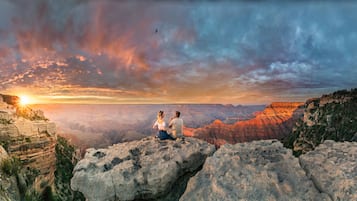A couple sitting on a rocky outcrop overlooking a canyon with a sunset in the background.