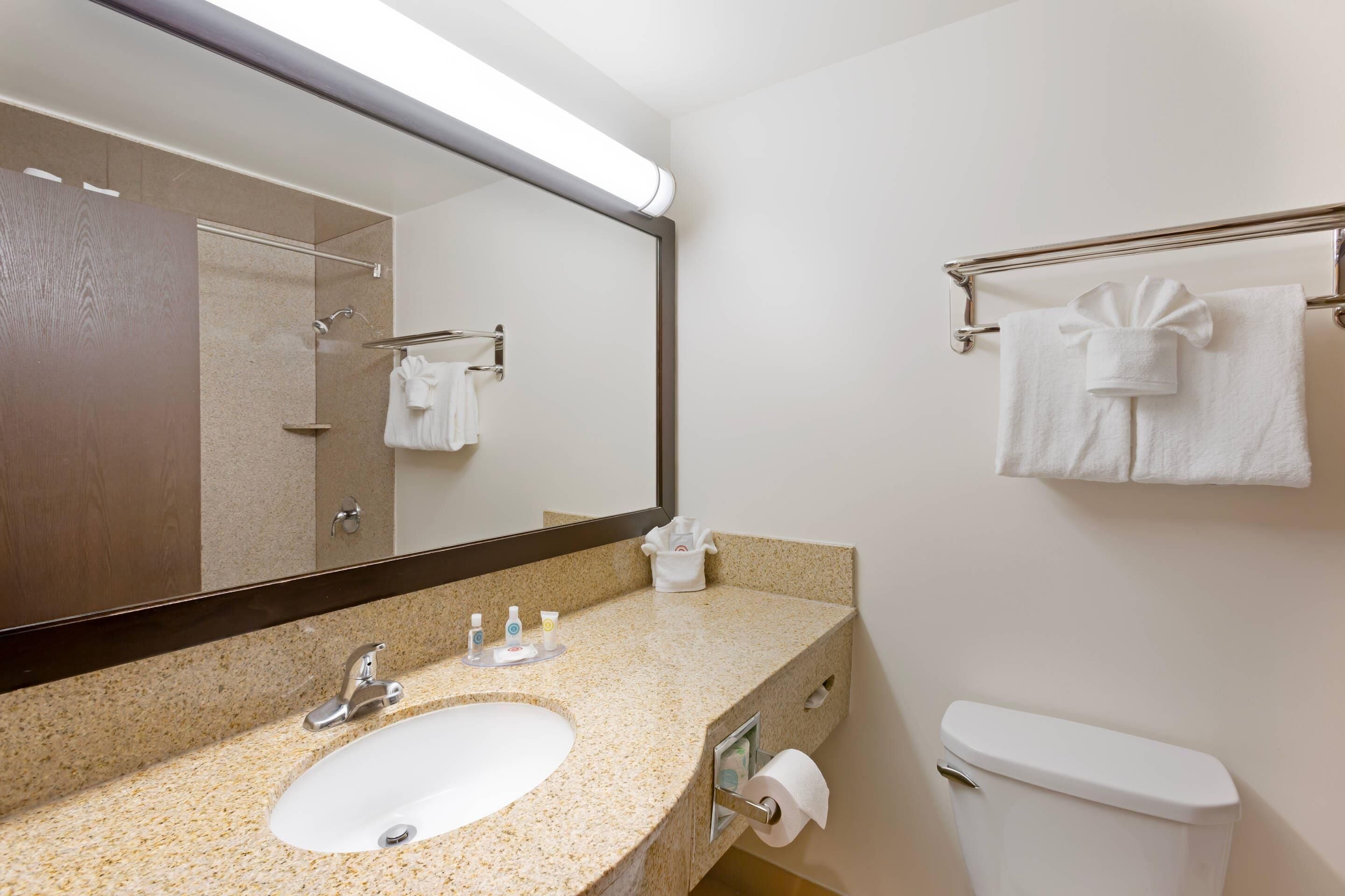 A hotel bathroom with a sink, mirror, towel rack, toilet, and a towel dispenser.