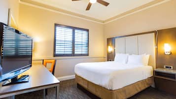 Habitación de hotel con cama, escritorio con televisor, ventana con persianas, ventilador de techo y luces fijadas a la pared.
