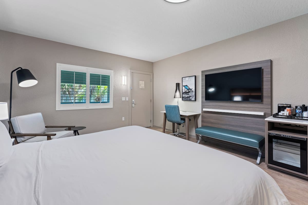 Ein Hotelzimmer mit einem großen Bett, einem Schreibtisch mit Stuhl, einem Fernseher und einem Fenster mit Jalousien.