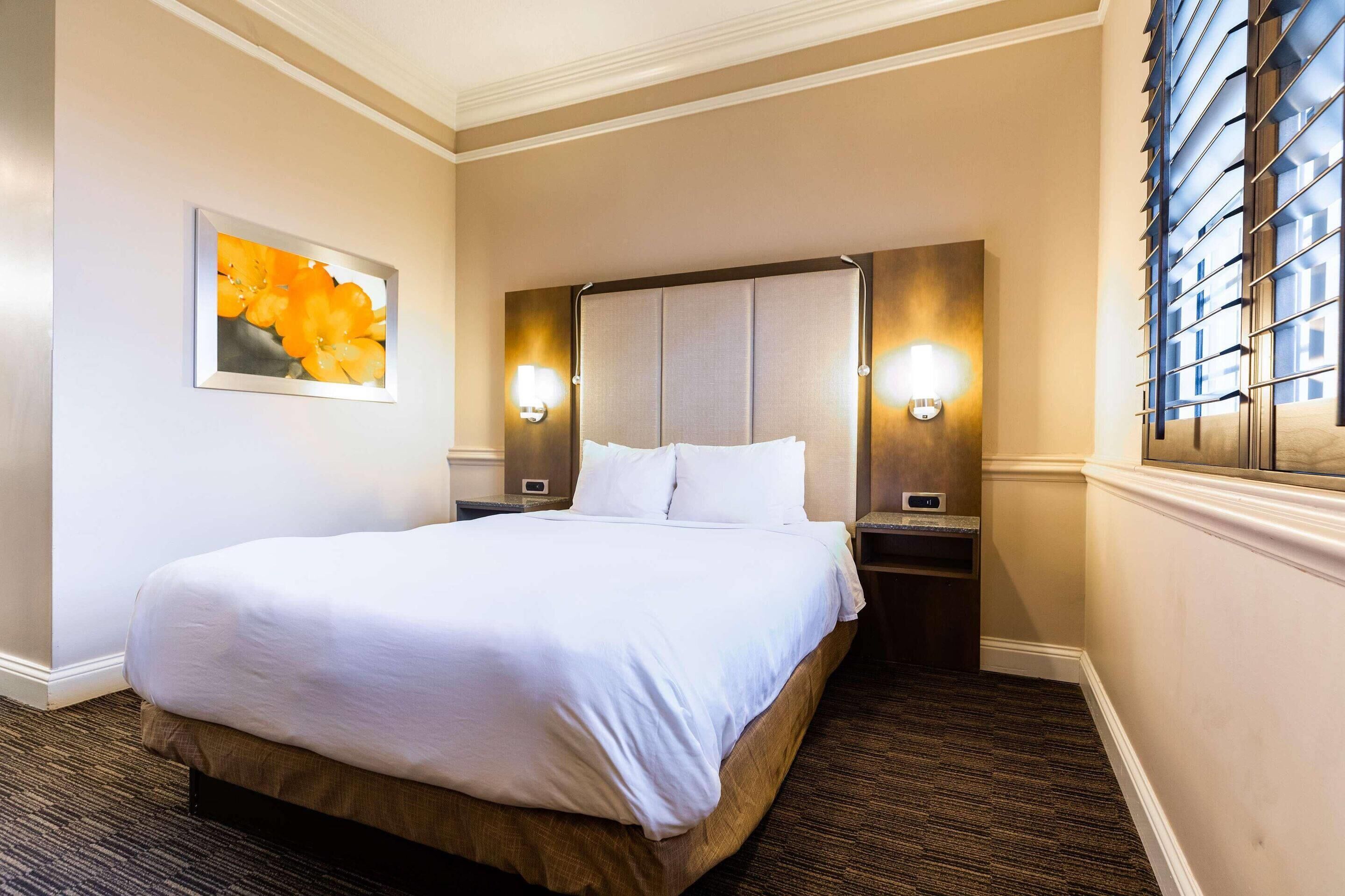 Habitación de hotel con una cama grande, dos mesitas de noche, un cabecero, una lámpara de pared y una ventana con persianas.