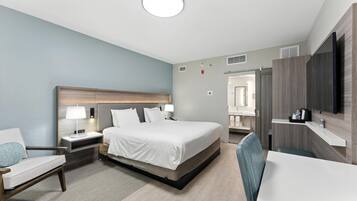 A modern hotel room with a large bed, bedside tables, a desk, a chair, a TV, and a bathroom with a mirror and sink.