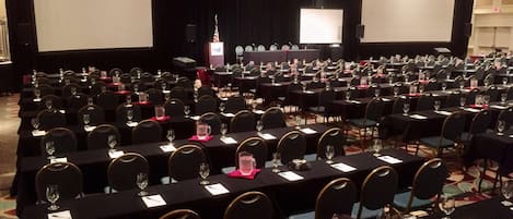 A conference room with rows of tables, chairs, and a stage with screens.