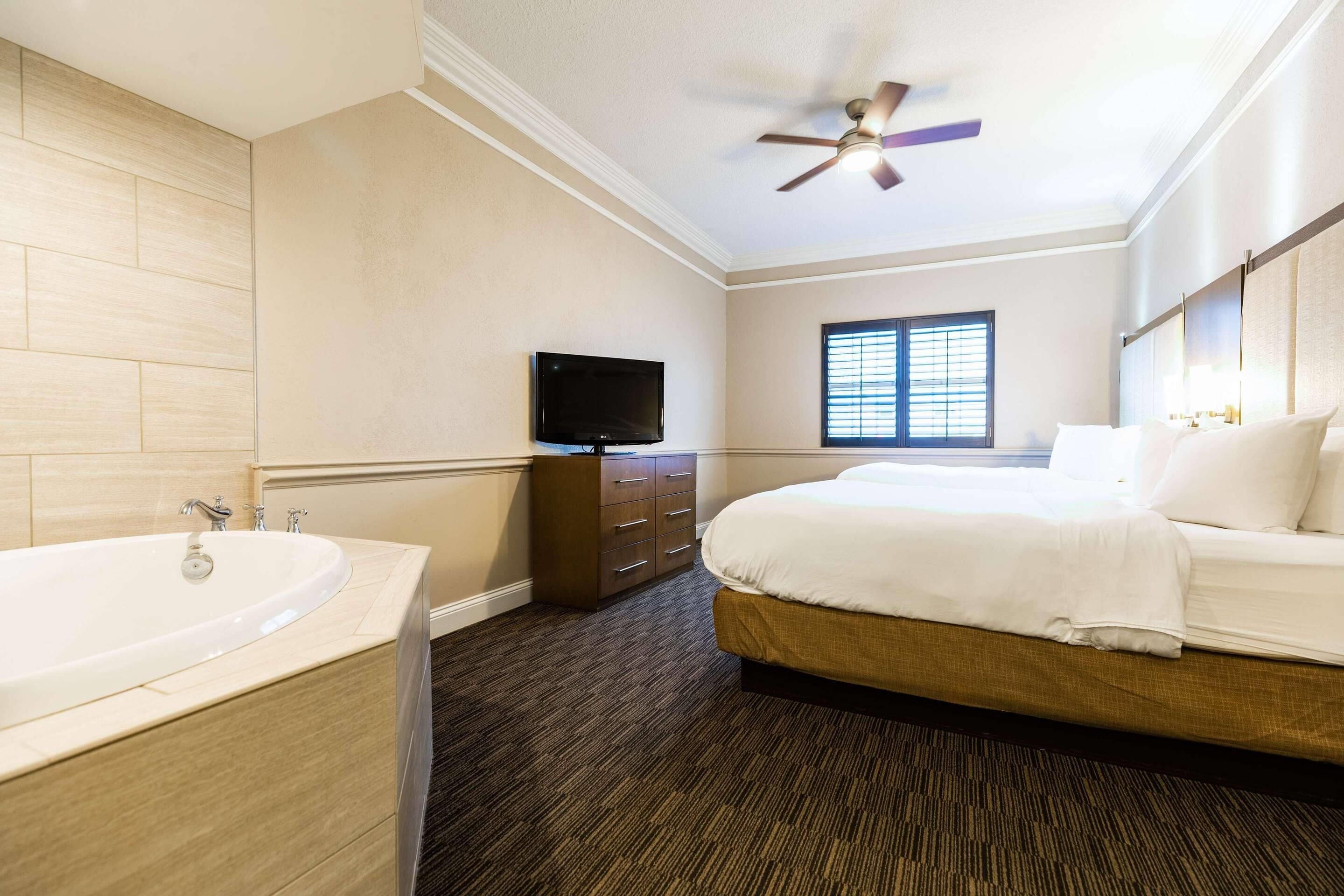 A hotel room with a bed, a television, a window with blinds, a ceiling fan, and a bathroom with a sink and mirror.