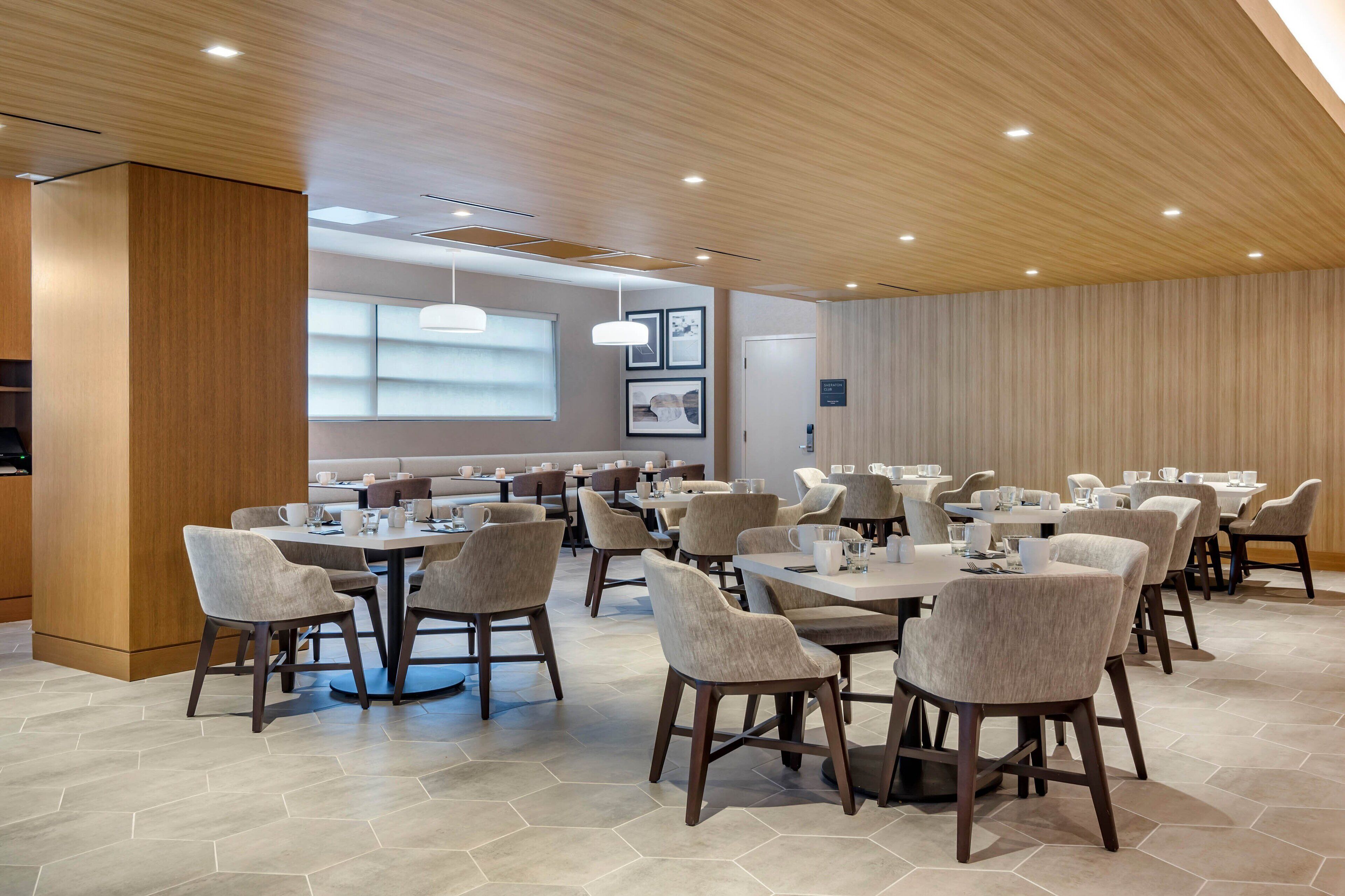 Een moderne eetruimte met meerdere tafels en stoelen, een houten plafond en een groot raam met jaloezieën.