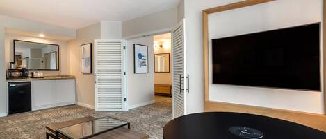 A modern hotel room with a kitchen area, a dining table, a flat-screen TV, and a large mirror.
