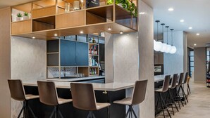 A modern bar area with high stools and a counter, featuring a wooden shelf with plants and bottles.