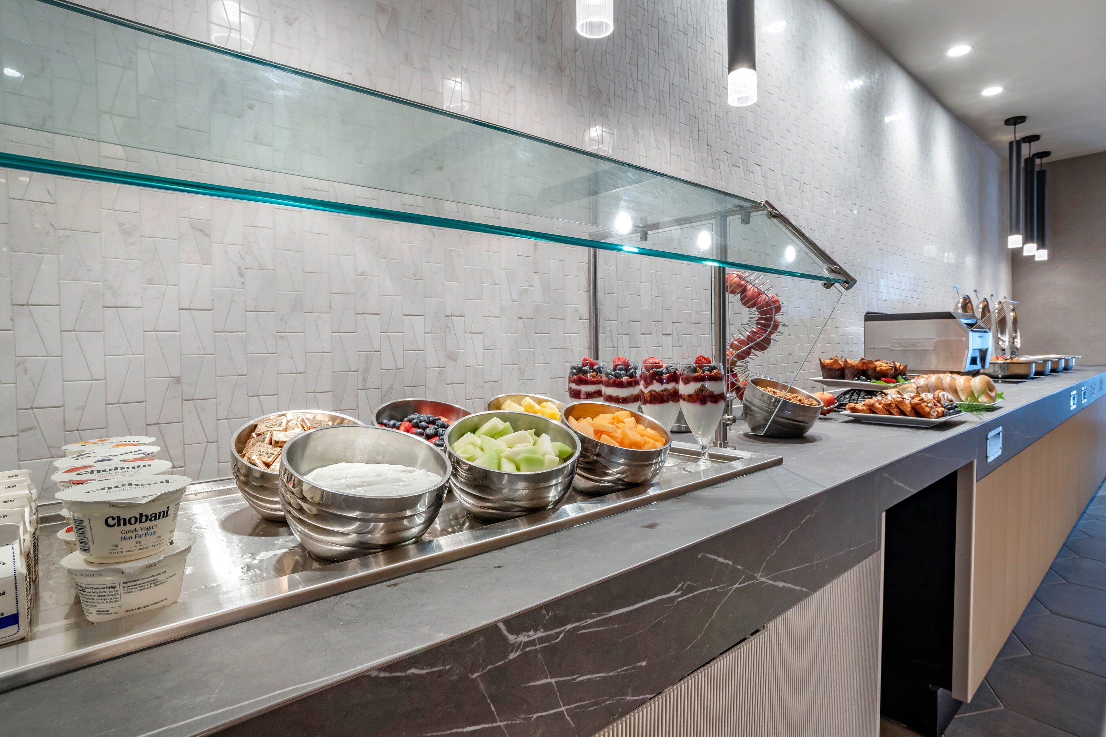 Zona de bufé con diferentes alimentos y recipientes, incluyendo tazones de frutas, yogur y otros bocadillos.