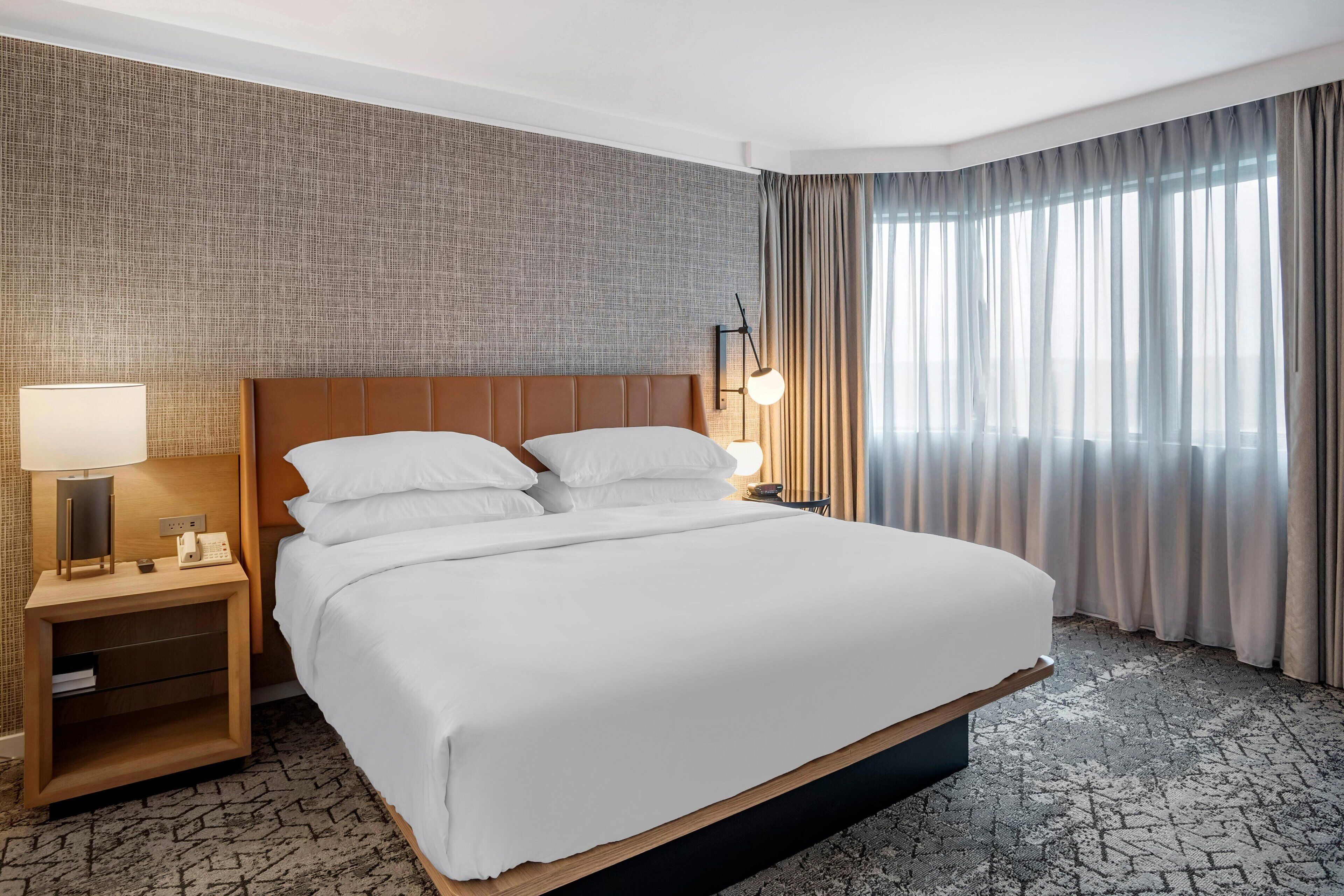 A hotel room with a large bed, a nightstand with a lamp, a wall-mounted phone, and a window with curtains.