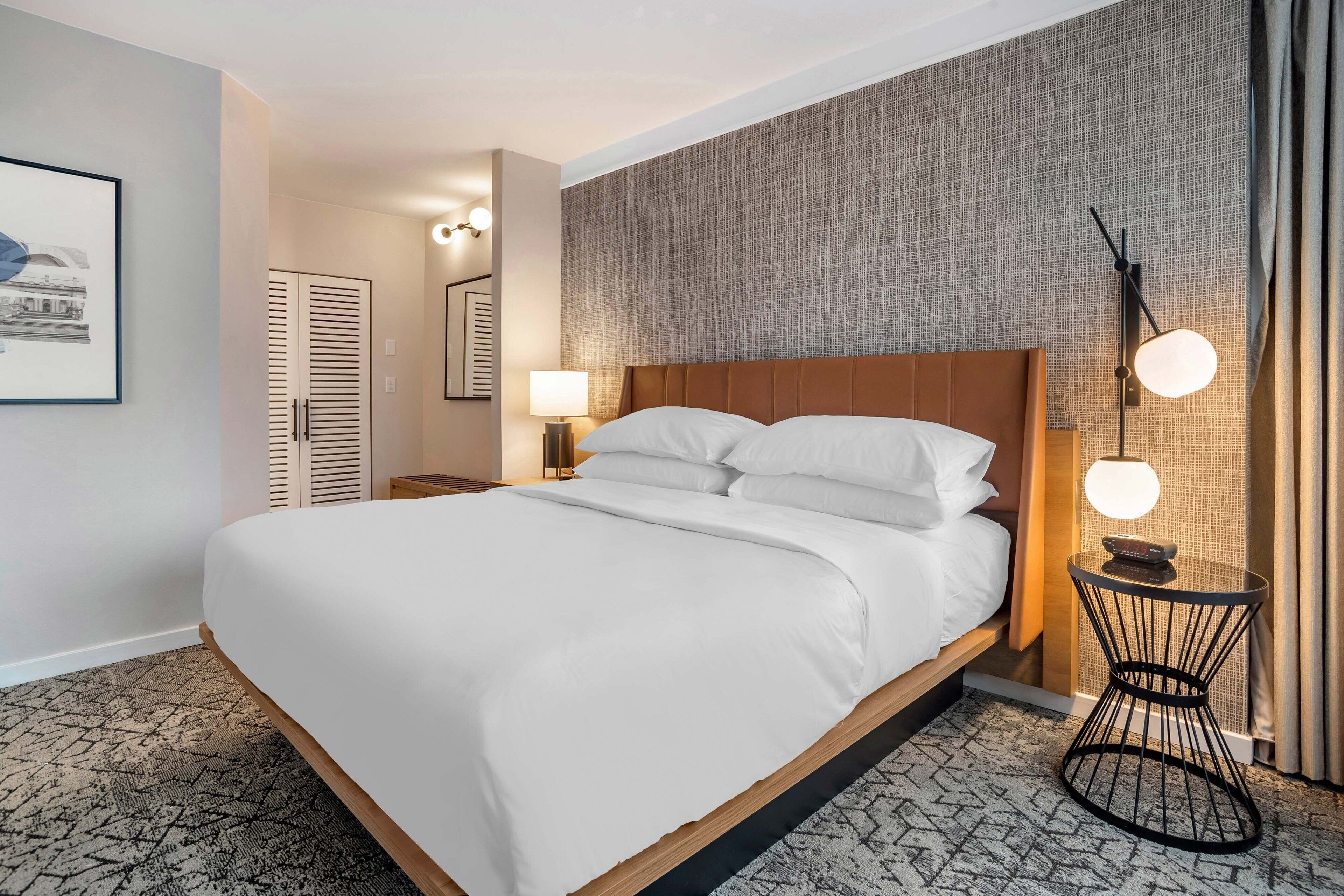 Une chambre d’hôtel avec un grand lit, une table de chevet avec une lampe, une horloge murale et un tableau encadré accroché au mur.