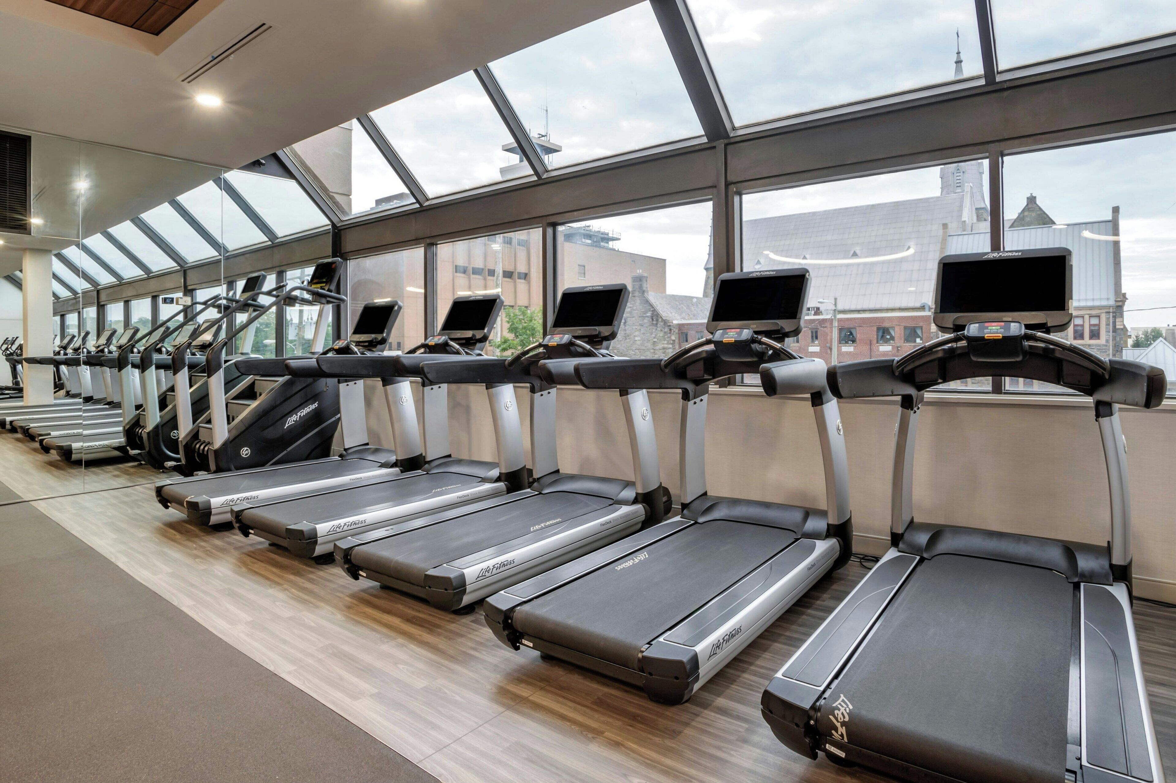 A row of treadmills in a gym with large windows and a view of buildings outside.