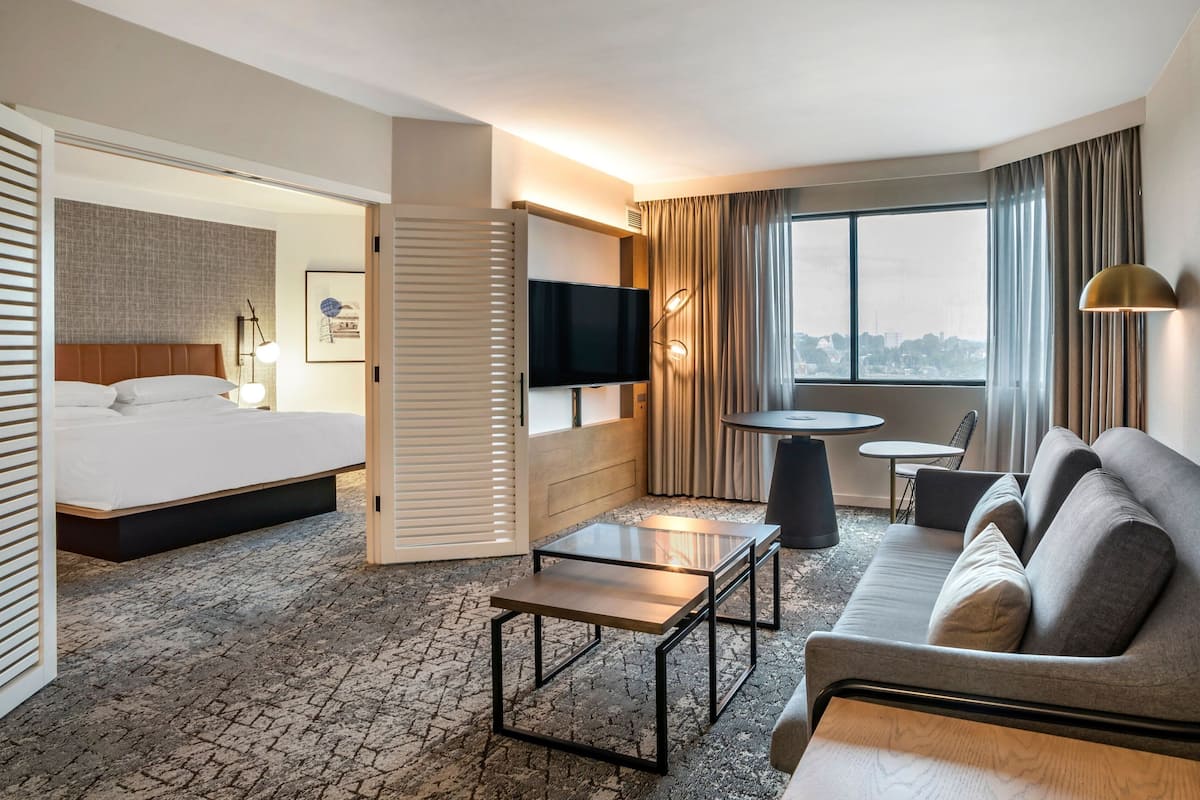 Een hotelkamer met een bed, een bank, een televisie, een kleine tafel en uitzicht op de stad door grote ramen.