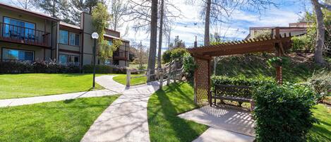 Un espacio exterior bien cuidado con un sendero de concreto, una pérgola de madera y un edificio con varias ventanas y balcones.