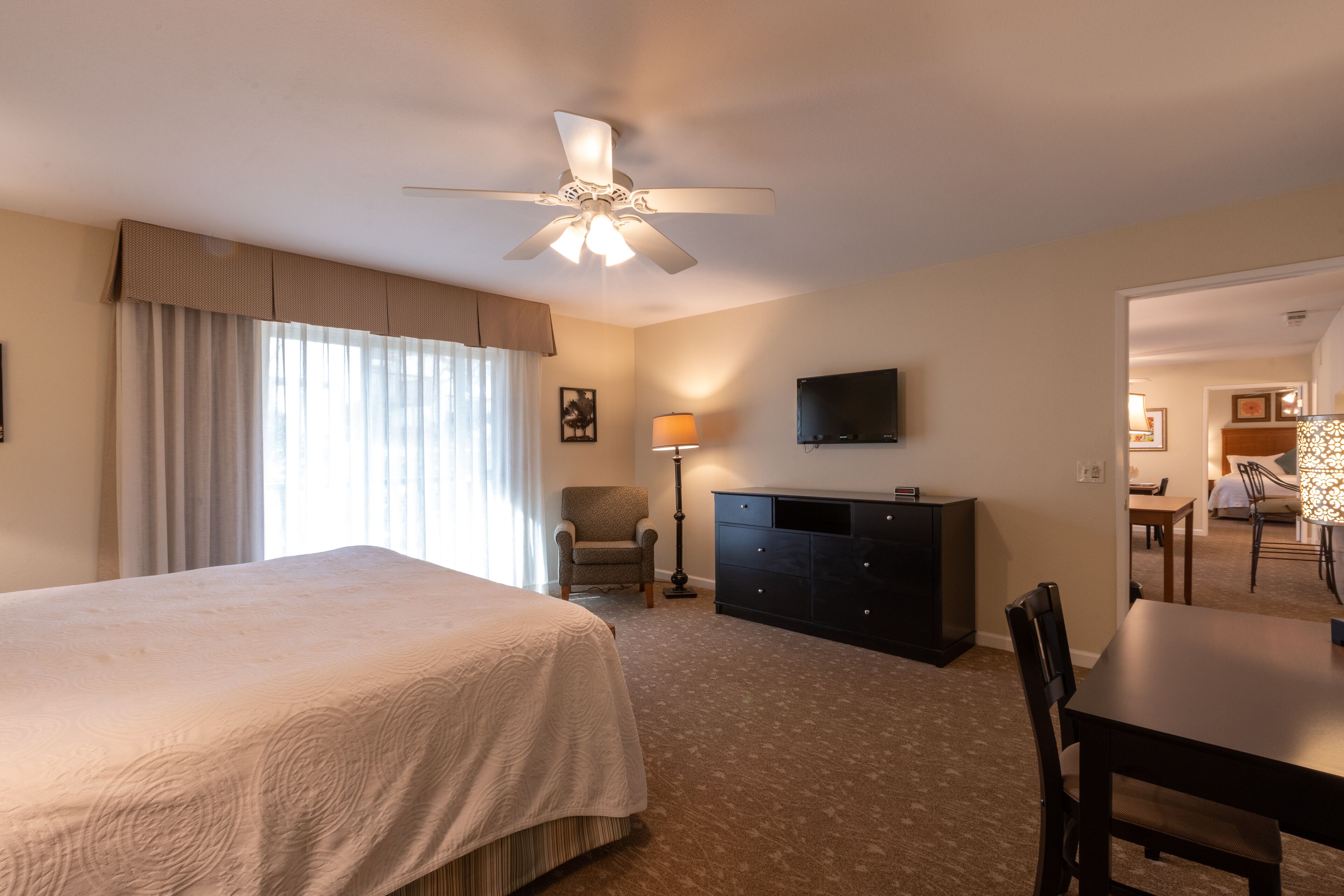 Ein Hotelzimmer mit Bett, Schreibtisch, Stuhl, Fernseher und Deckenventilator.