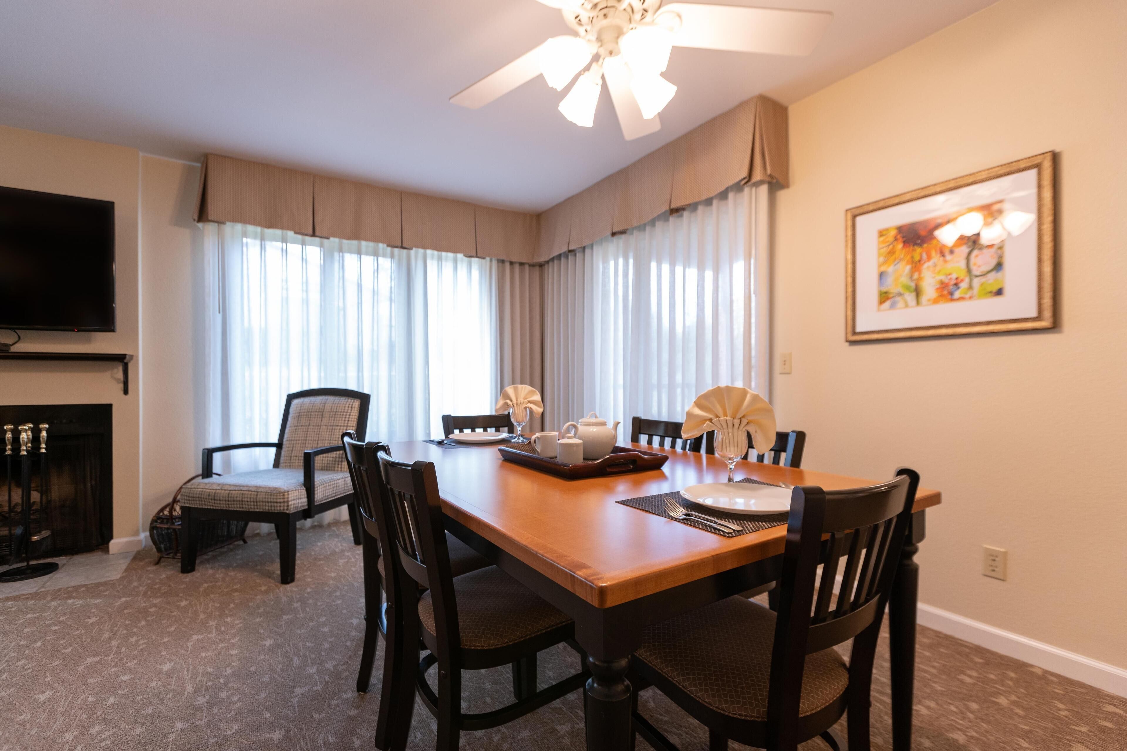 A dining room with a wooden table, four chairs, a ceiling fan, a fireplace, and a television on the wall.