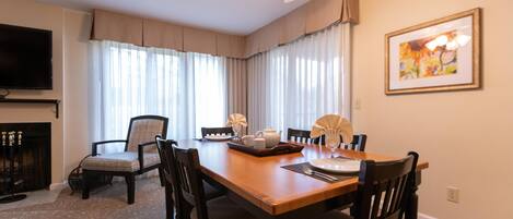 A dining room with a wooden table, four chairs, a ceiling fan, a fireplace, and a television on the wall.