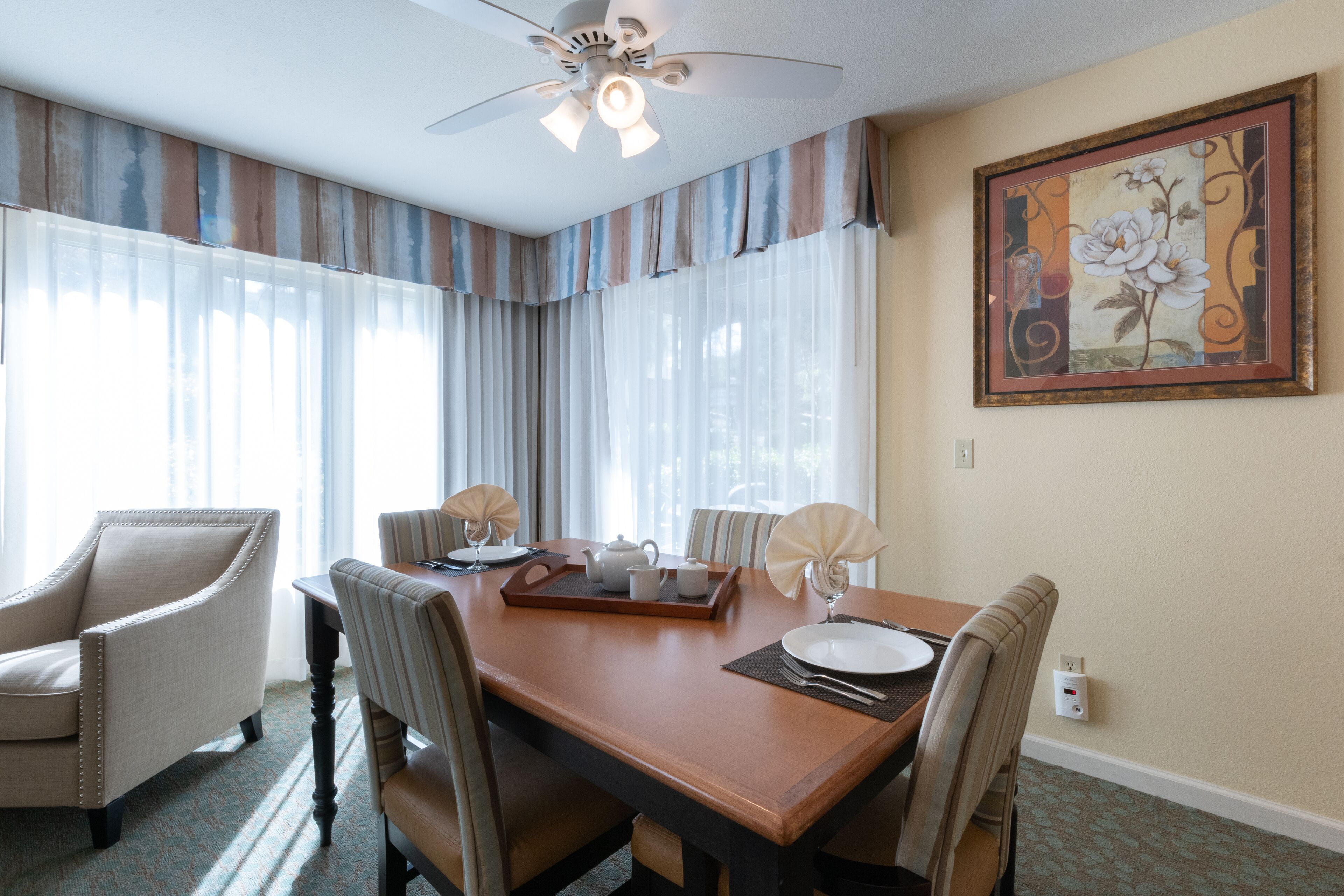 Un comedor con una mesa de madera, sillas, un ventilador de techo y un cuadro en la pared.