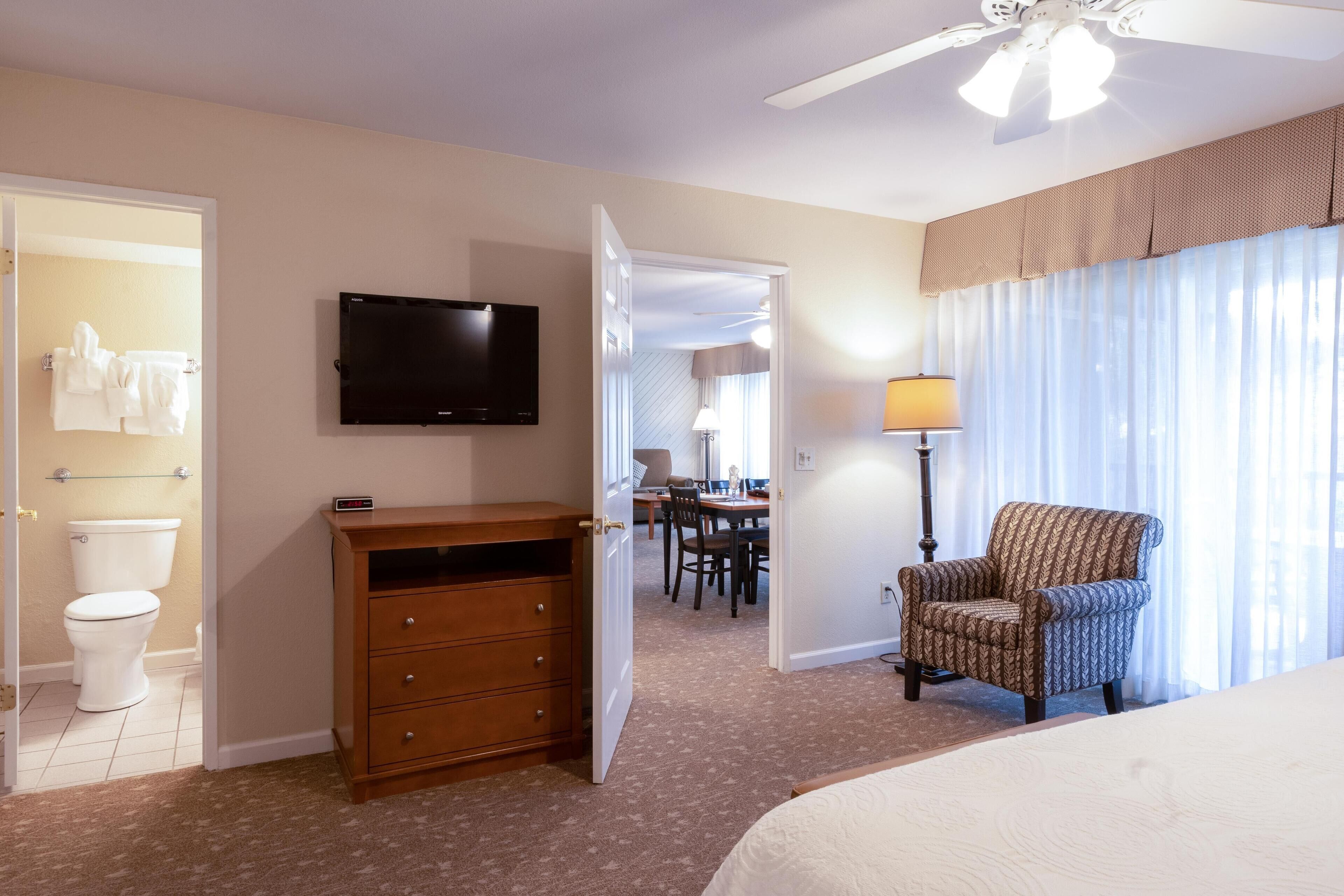 A hotel room with a bed, a television, a wooden dresser, a chair, a desk with a lamp, and a bathroom with a toilet and towel rack.