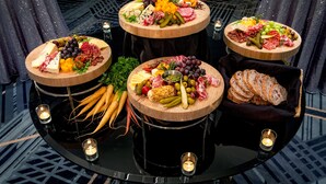 A round table with three wooden platters of assorted cheeses, meats, and fruits, surrounded by candles.