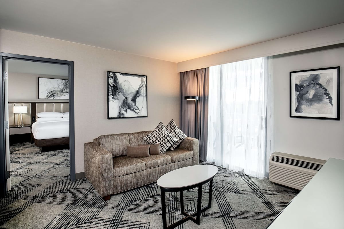 Une chambre d’hôtel avec un canapé, une petite table et un lit. Deux tableaux abstraits ornent les murs, et il y a une fenêtre avec des rideaux.