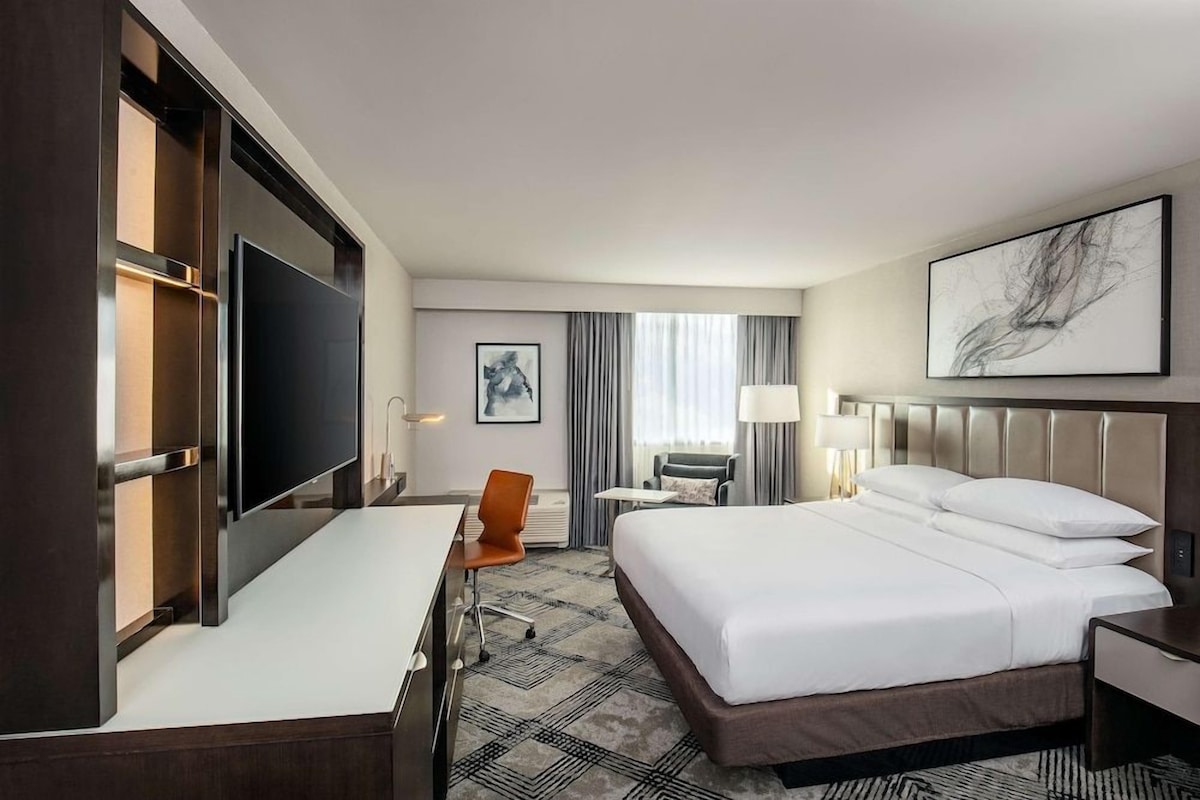 A hotel room with a bed, desk, chair, TV, and artwork on the walls.