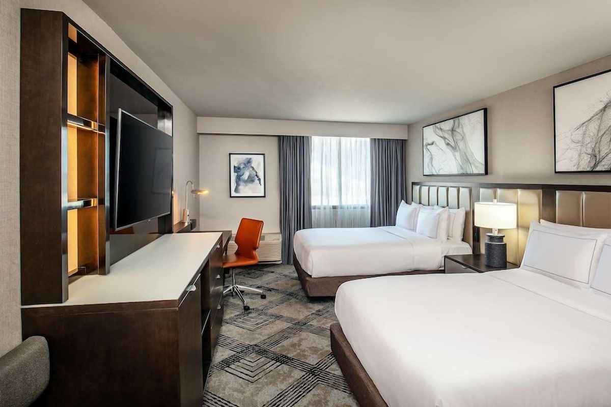A hotel room with two beds, a desk, a TV, and artwork on the walls.