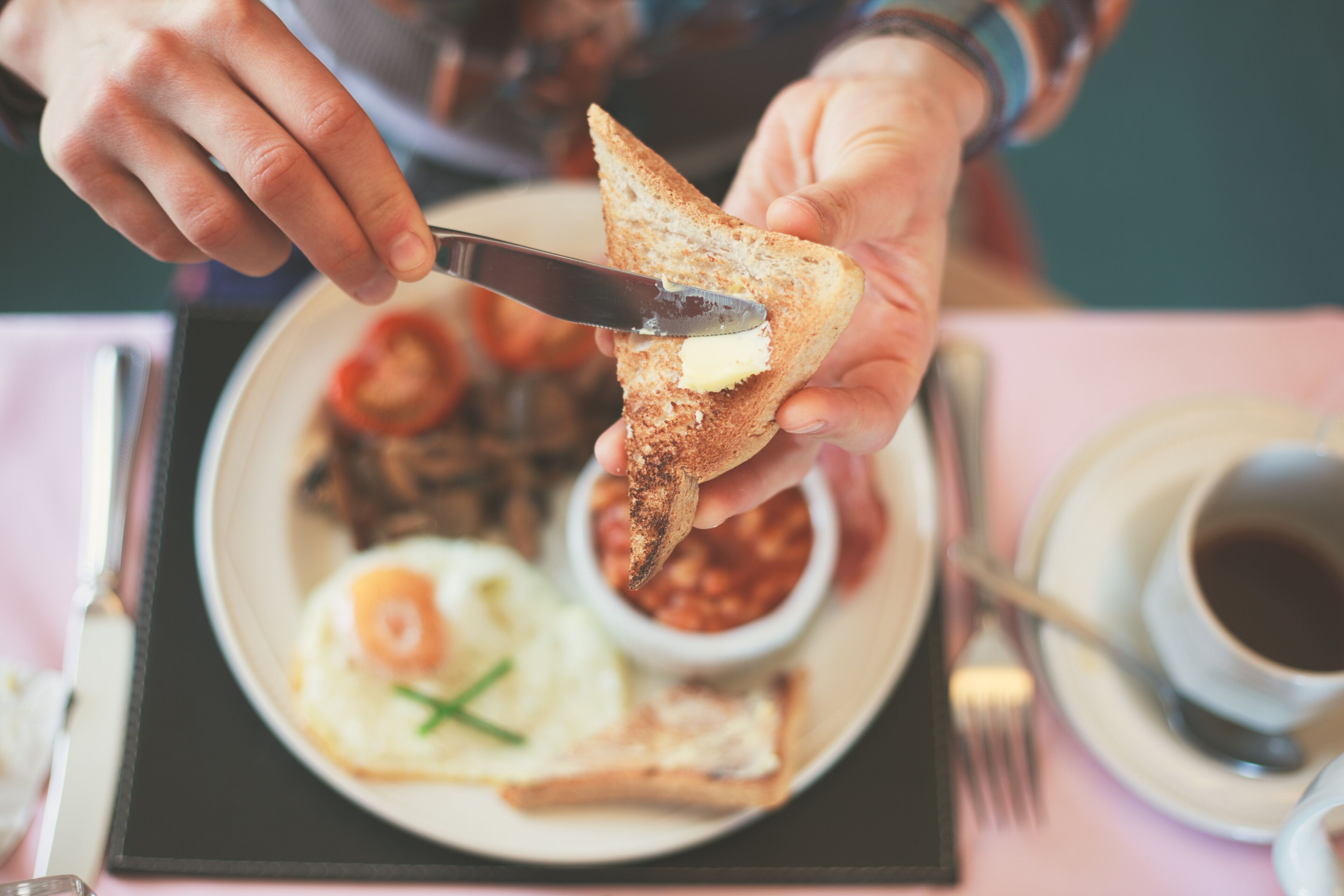 Une personne tartine du beurre sur une tranche de pain grillé, avec une assiette d’œufs et de haricots en arrière-plan.
