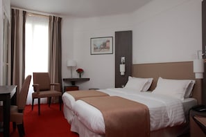 Hotellihuone, jossa on kaksi sänkyä, työpöytä, tuoli, pieni pöytä ja taulu seinällä.