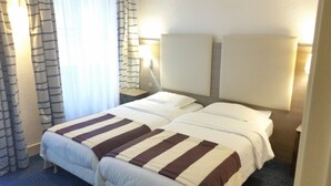 Quarto de hotel com duas camas, roupa de cama listrada, uma mesa de cabeceira e um abajur.