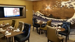 Uma área de refeições com mesas preparadas para quatro pessoas, cada uma com uma cadeira, uma televisão montada na parede e um grande mural de uma onda na parede atrás das mesas.