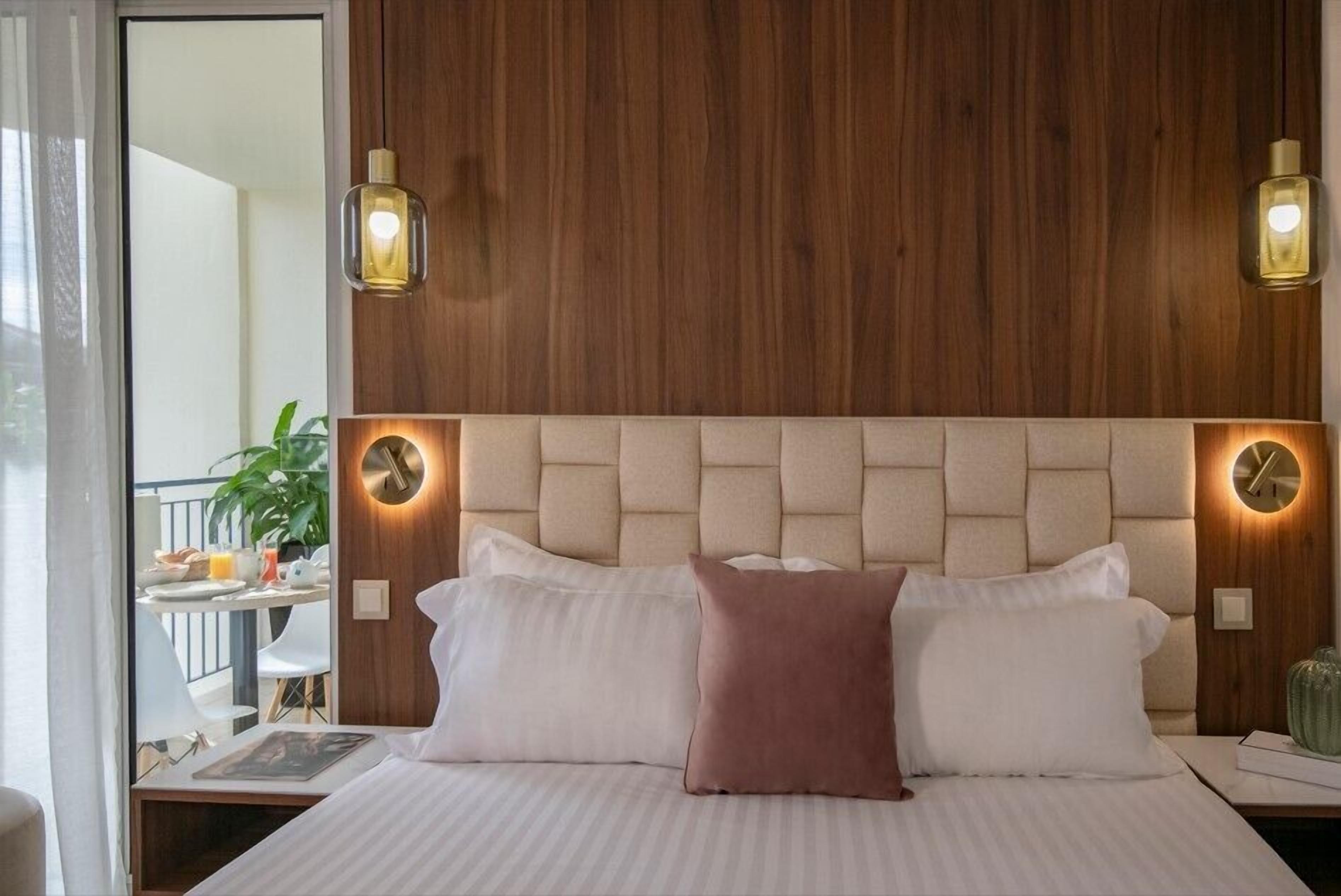 A hotel room with a wooden headboard, two bedside lamps, a bed with white and pink pillows, and a view of a balcony with a table and chairs.