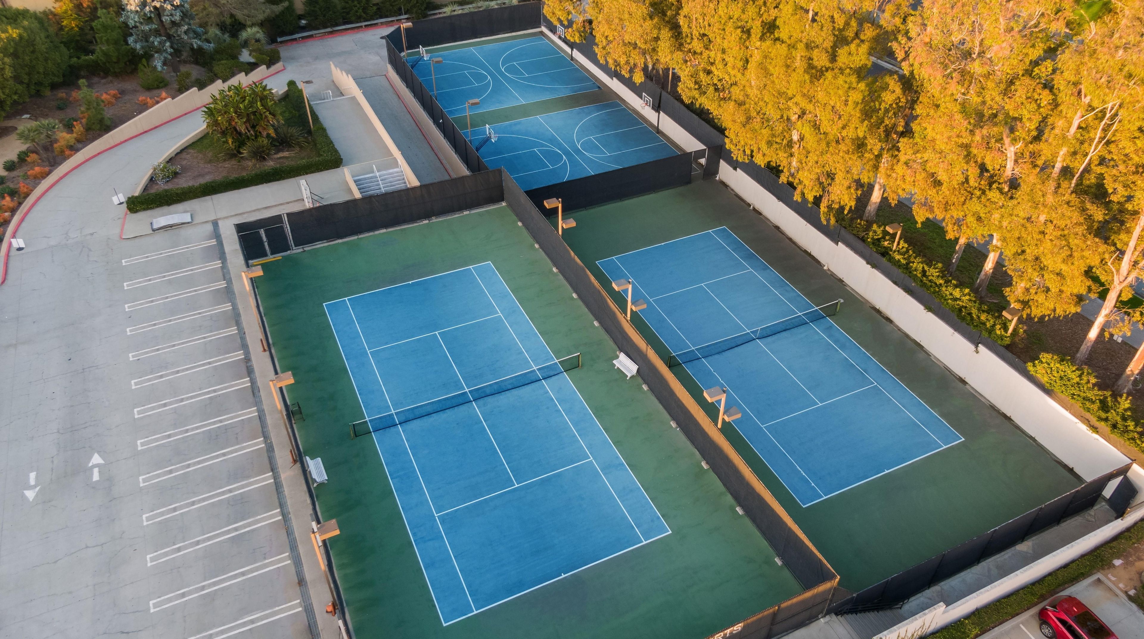 An aerial view of a multi-sport court with basketball and tennis courts, surrounded by trees and a parking area.