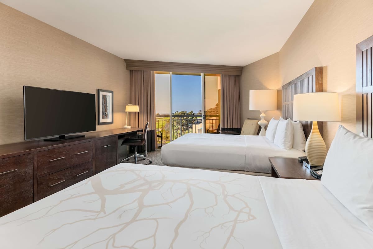 A hotel room with two beds, a TV, a desk, and a balcony with a view.