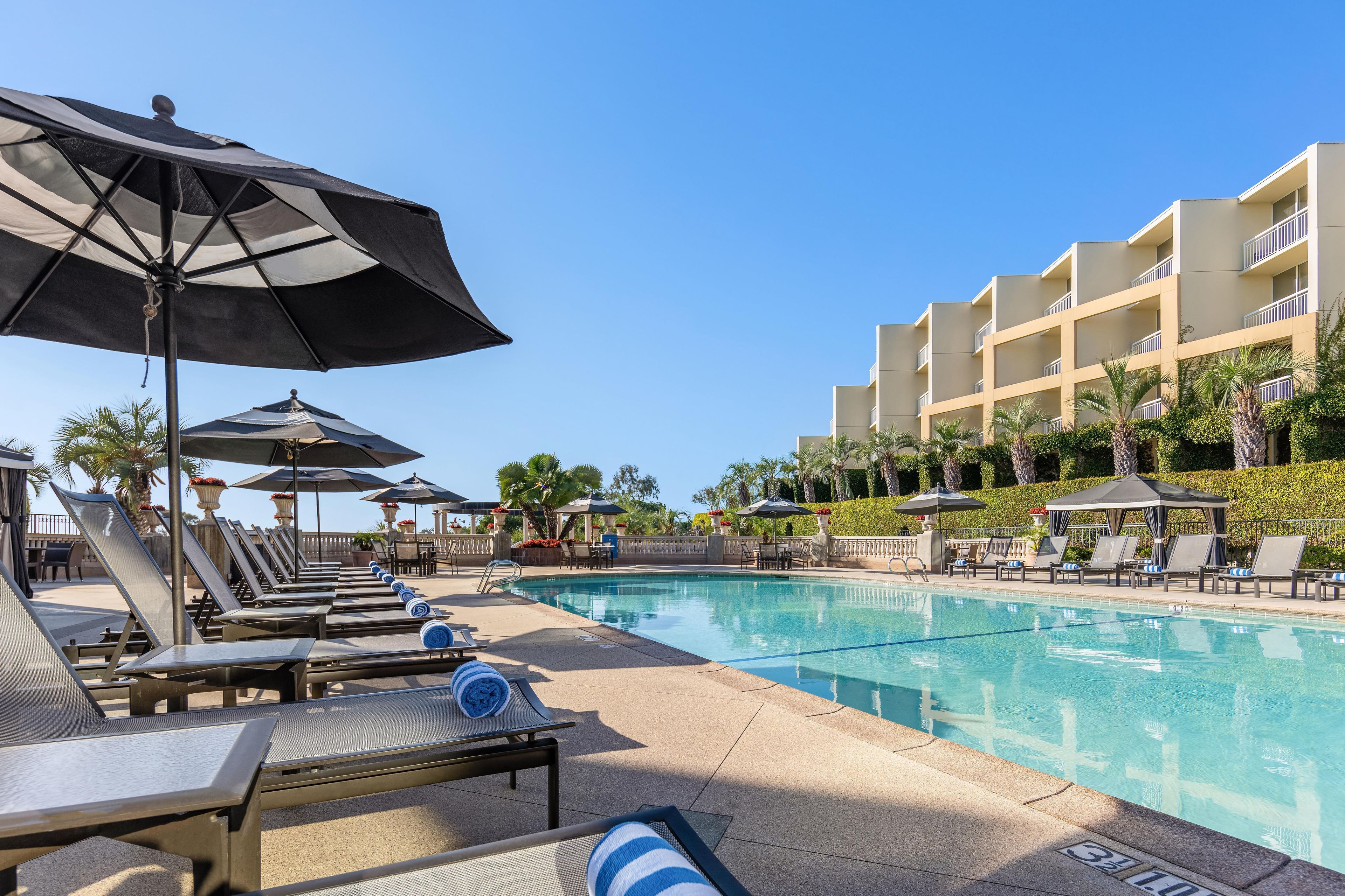 A poolside area with lounge chairs and umbrellas, a clear blue sky, and a building with balconies.